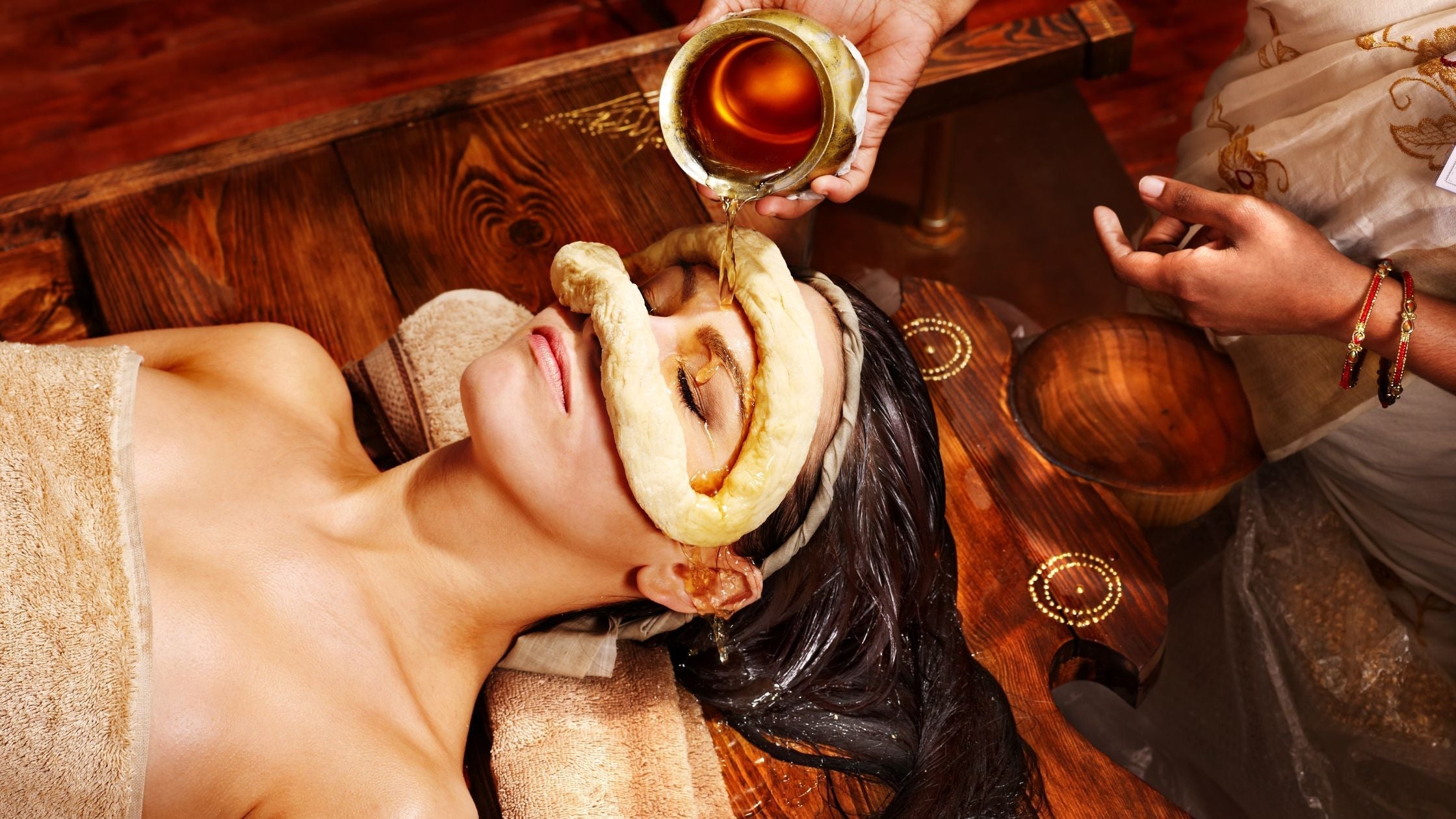 An Ayurvedic practitioner pours warm herbal oil into a dough dam placed on a woman's face during a holistic Panchakarma therapy session.