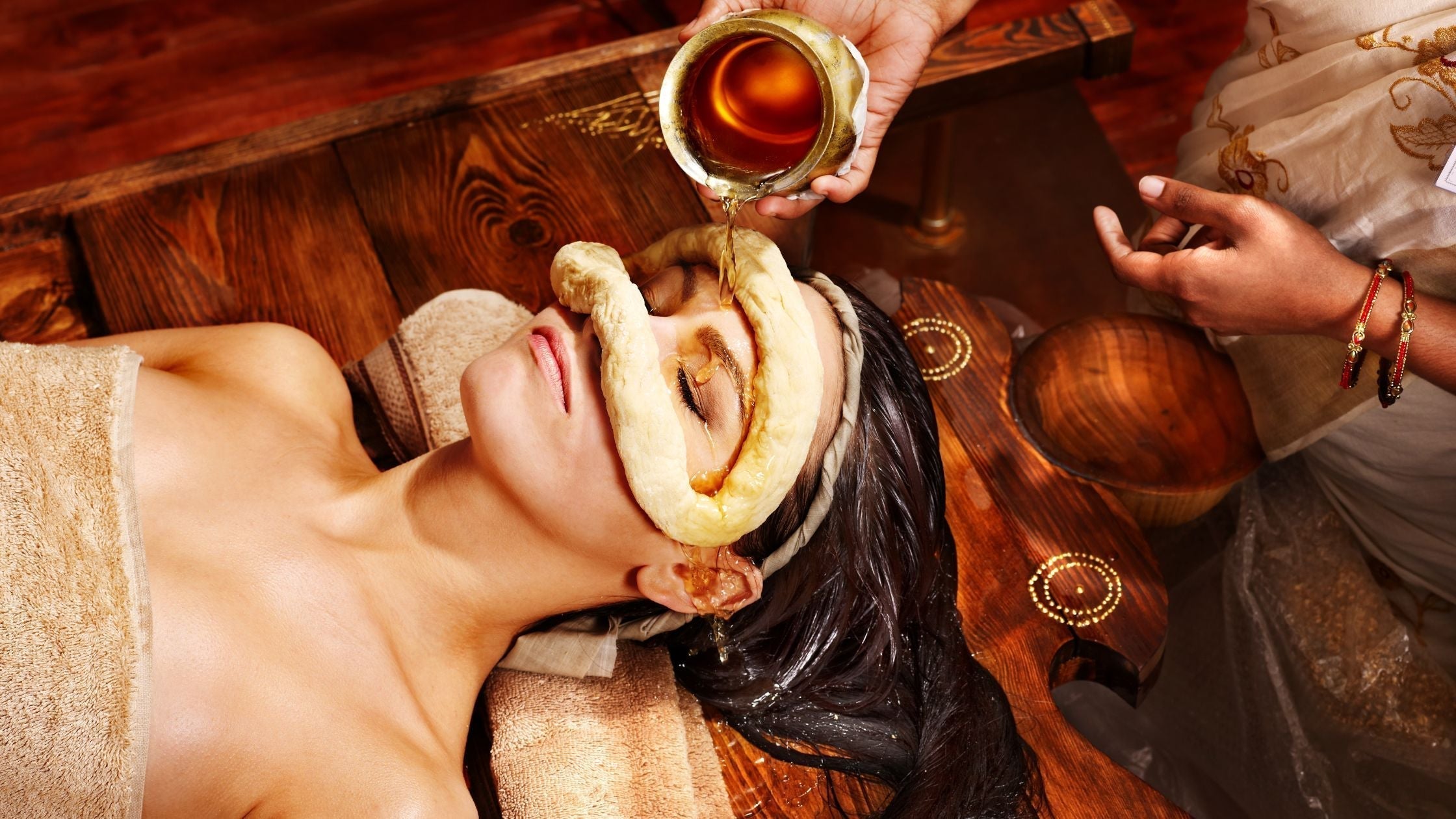 An Ayurvedic practitioner pours warm herbal oil into a dough dam placed on a woman's face during a holistic Panchakarma therapy session.